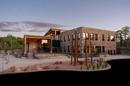 A modern two-story stone building with large windows, outdoor seating steps, and metal sculptures in the landscaped front area at dusk.