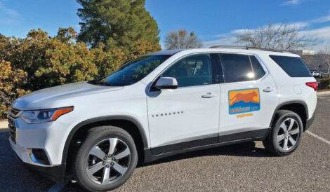A white Chevrolet Traverse SUV with a colorful logo on the front door is parked in a lot near trees under a clear sky.