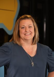 A woman with shoulder-length blonde hair, wearing a dark blouse and a silver necklace, smiles at the camera with a yellow and blue background behind her.