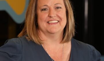 A woman with shoulder-length blonde hair, wearing a dark blouse and a silver necklace, smiles at the camera with a yellow and blue background behind her.