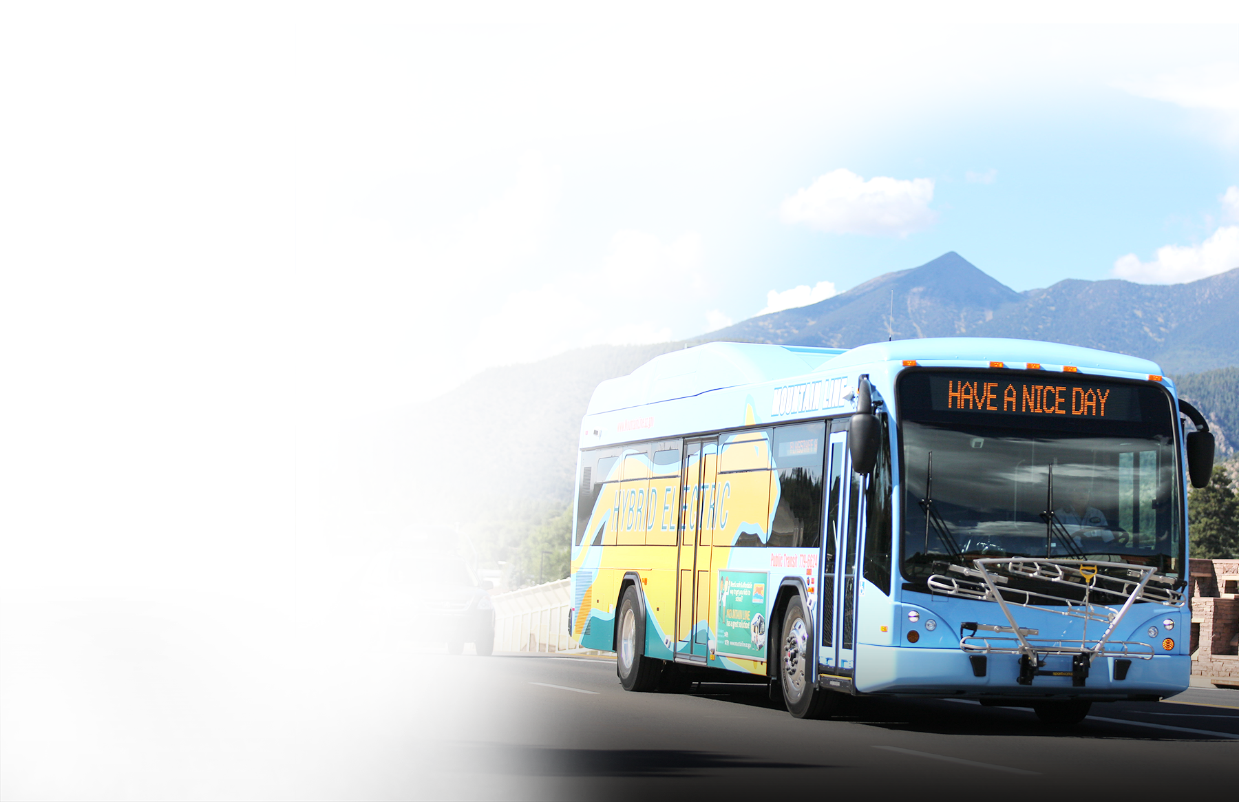 A light blue Mountain Line hybrid electric bus, its sign reading “Have a nice day,” driving past mountains.