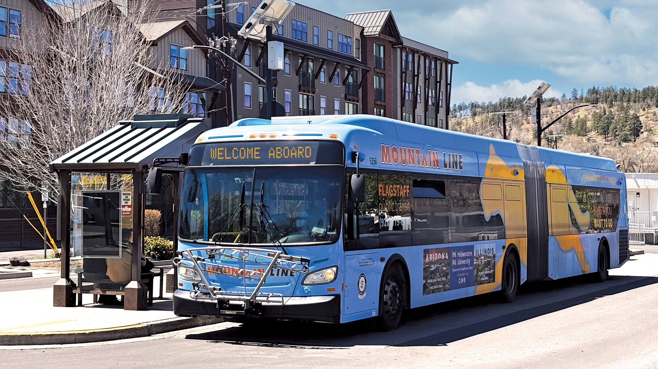 A Mountain Line articulated bus at a downtown Flagstaff stop, its sign reading Welcome Aboard.